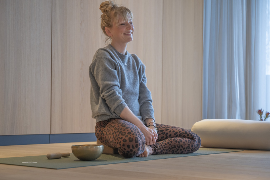 Frau sitzt im Schneidersitz auf einer Yogamatte mit Klangschale am Fenster im Seegut Zeppelin