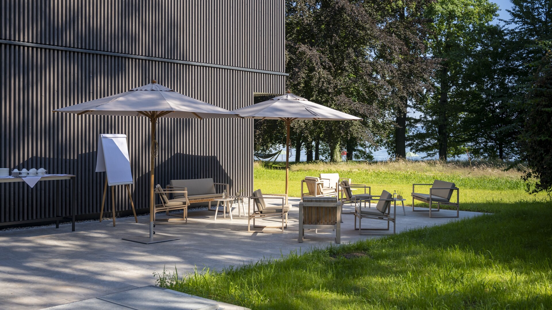 Terrasse am Buchensaal des Seegut Zeppelin mit Sonnenschirmen, Loungemöbeln und Flipchart vor Wiese und Bäumen