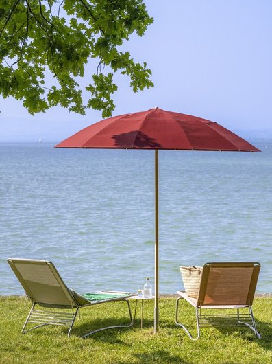 Roter Sonnenschirm und zwei Liegestühle am Ufer des Bodensees im Sommer
