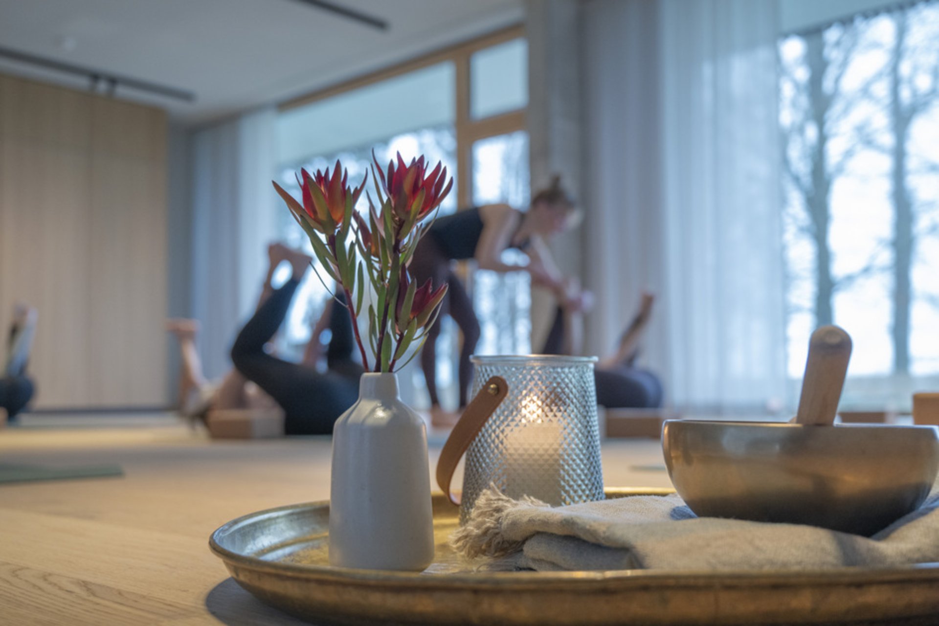 Klangschale, Kerze und Blumenvase auf einem Tablett, im Hintergrund Menschen beim Yoga im Seegut Zeppelin